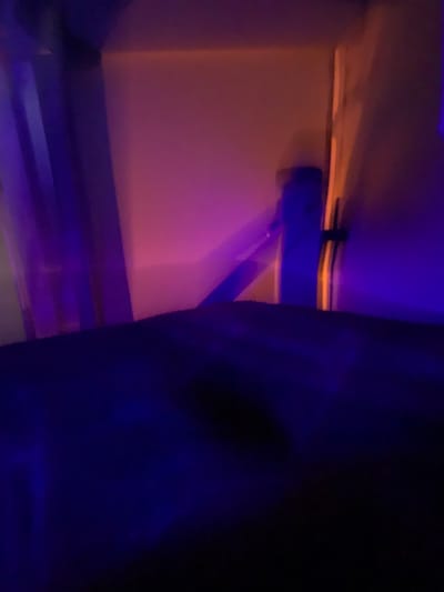 The image displays a blurred, dimly lit interior space, possibly a room or shelf. A peach-colored wall contrasts with a shadowy figure in the foreground. A beam or shelf-like structure spans the width of the frame, highlighted by blue and orange lighting. The overall atmosphere is dark and unclear due to motion blur and low lighting.