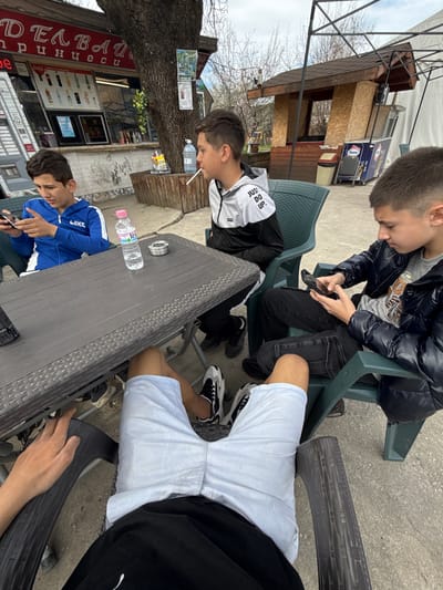 Four young males are seated around a gray outdoor table, engaged in activities, possibly at a café. One individual in a blue jacket is focused on a mobile phone. Another youth, wearing a white and black jacket, has what appears to be a straw in his mouth. The third individual, adorned in a dark puffer jacket, is also using a mobile phone. A fourth person, wearing a light-colored shirt, is partially visible. The table holds an ashtray and a water bottle. In the background, there is a small building with signage, including text in Cyrillic and Roman alphabets, and advertisements for beverages such as Coca-Cola. Based on the signage ("ДЕЛВАЙ принцеси"), the location is likely in Zlatna Panega, Bulgaria.