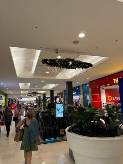 The image displays the interior of a brightly lit shopping mall, adorned with festive decorations. A large, inverted Christmas tree, decorated with white lights and a gold ribbon, hangs from the high ceiling, complemented by strings of white fairy lights draped across the space.
Several shoppers are visible, primarily in the lower portion of the image. In the foreground, a woman with light, curly hair wears a denim jacket over a green dress or skirt and carries a brown bag. Other individuals are seen walking or standing further down the concourse.
In the mid-ground, a black, futuristic massage chair with blue illumination is on display next to a large potted plant housed in a white cylindrical planter. A digital screen nearby shows the text "Feel Good." The mall features modern architecture with white walls, high ceilings, and recessed circular lights, along with rectangular illuminated panels.
Shopfronts line both sides of the mall. A prominent red and blue sign is partially visible, displaying "THE" in blue and an orange "SAVE" logo, indicating a retail outlet. Other blurred shop displays are also present. The scene captures typical shopping activity during what appears to be the holiday season, with people browsing and moving through the well-lit indoor environment. Given the context, this is likely a shopping center in Narre Warren, Australia.