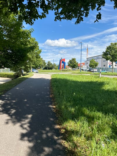 The image displays an outdoor scene at a traffic roundabout in Schongau, Germany, specifically at the intersection of B17 (Altenstadter Straße) and Münzstraße/Bahnhofstraße. The scene is illuminated by bright sunlight under a clear blue sky with scattered white cumulus clouds and some wispy cirrus streaks.

In the foreground, a dark gray asphalt path extends from the lower left towards the center, partially covered by the dappled shadows of overhead tree foliage, which frames the upper portion of the image. To the right of the path is a vibrant green grassy verge.

The midground is dominated by the central roundabout, which features a prominent, abstract art sculpture known as "Schongauer Brücke." The sculpture consists of multiple angular planes in red and blue, with some metallic gray components. The center of the roundabout is a manicured grassy area. Several green deciduous trees line the perimeter of the roundabout and the adjacent roads. A light-colored sedan is observed on the roundabout's paved road on the left. On the right side of the roundabout, near buildings, a dark-colored vehicle and a light blue vehicle are visible. Two tall, blue-grey streetlights stand upright, one behind the sculpture and another further right. Standard German traffic signs, including a yield sign and a bicycle path sign, are positioned on the right.

In the background, multi-story, light-colored buildings are visible on the right, suggesting residential or commercial structures. A tall yellow construction crane is prominently positioned behind these buildings. No human figures are present within the depicted scene.