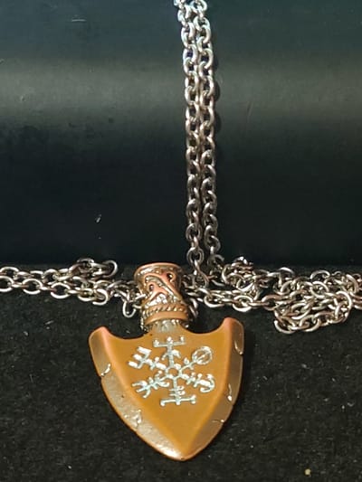 The image shows a close-up, slightly blurry photograph of a silver-colored metal chain necklace with a pendant. The pendant is shaped like an arrowhead and is a coppery-brown color with intricate silver-colored etchings or symbols on its surface. The necklace is resting on a dark, textured surface, likely fabric. The background is a dark, out-of-focus area, possibly a black surface or cloth. There are no people or actions visible in the image. The location in Sokolohirsk, Ukraine is not depicted or suggested in the visual information.