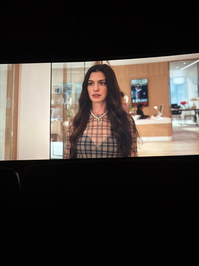 A medium shot shows a woman with long brown hair and a pearl necklace wearing a plaid shirt. She is positioned in front of an office space with a large window and various furnishings. This office space is located in the city of Rapperswil-Jona, Switzerland.