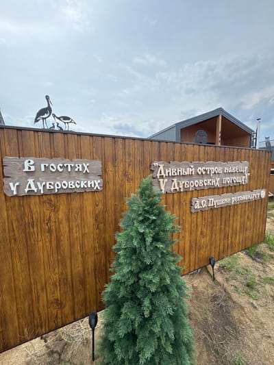 The image depicts a wooden fence with two signs and decorative metal bird silhouettes on top. The left sign reads "В ГОСТЯХ У ДУБРОВСКИХ" (Visiting the Dubrovskys). The right sign contains text in quotes: "“Дивный остров навещу У Дубровских погощу!”" (I will visit the wondrous island, I will visit the Dubrovskys!). Below this, another line reads "А.С. Пушкин рекомендует" (A.S. Pushkin recommends), with a smiling emoji. A small evergreen tree is planted in front of the fence. In the background, a modern building with a gabled roof is visible. The scene appears to be outdoors, likely in a rural or semi-rural setting. The location is identified as Khuzhir, Russia.