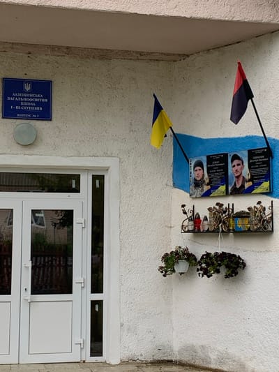 An exterior wall of the Lazeshchyna General Educational School, Building No. 2, in Lazeshchyna, Ukraine, is depicted. The wall is white and textured. To the left, a white double-door entrance with glass panes is visible. Above the doors, a blue sign with the school's name and building number in Ukrainian is mounted. To the right of the doors, a memorial display is present on a blue and white painted section of the wall. Two flags on poles extend from the wall: a blue and yellow Ukrainian national flag on the left, and a red and black flag on the right. Below these, two dark-framed memorial plaques are affixed, each displaying a photograph of a young man and biographical information in Ukrainian, identifying them as Mykola Romanovych Yatsko (born 20.06.1993, died 06.02.2023) and Dmytro Ivanovych Havryliuk (born 26.11.1982, died 27.08.2022). Beneath the plaques, a black wrought-iron shelf with decorative elements holds two red and one white votive candles in glass containers, alongside a small yellow and blue decorative object. Below the shelf, two white hanging planters containing dark-flowering plants are suspended. The ground immediately in front of the wall appears to be paved.