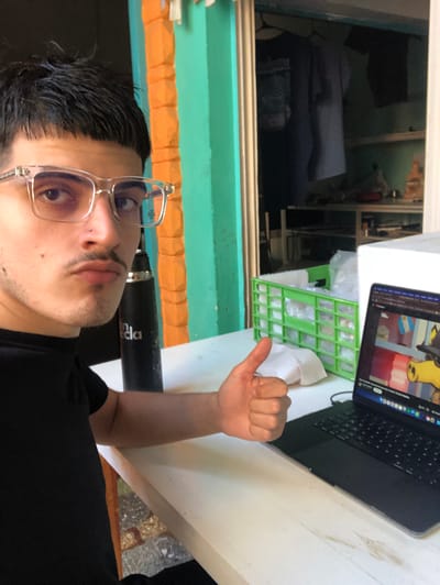 A selfie of a person with short, dark hair and clear-framed glasses. The person is wearing a black shirt and has a piercing in their nostril. They are holding up a thumb. In the background, a laptop is open with a cartoon image on the screen. A green egg crate with white packaging inside sits next to the laptop. A black water bottle is also visible. A window behind the person shows a room with a dark bed and some shelves. The wall around the window is painted in horizontal stripes of orange, light blue, and green. The location is likely an indoor setting in Beccar, Argentina, based on the flag of Argentina displayed on the screen of the laptop.