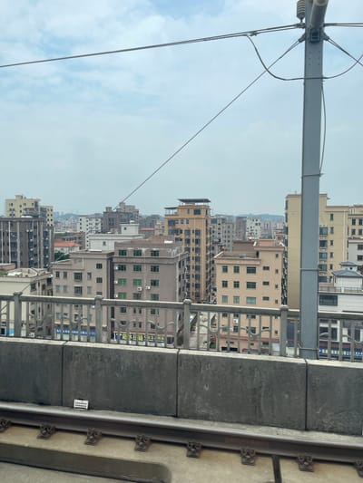 This image captures a view of a dense urban landscape, likely in Dongguan, China, from a slightly elevated perspective, possibly from a train or a tall building. The scene is dominated by numerous apartment buildings of varying heights and architectural styles, packed closely together. The buildings are predominantly light-colored, with visible windows, air conditioning units, and balconies. Some buildings feature different colors, adding a subtle visual variation to the cityscape.
In the foreground, a metal railing and what appears to be a concrete barrier suggest a structure like a railway bridge or elevated walkway. Overhead, electrical wires are visible, indicating the presence of transportation infrastructure. The sky is overcast with some scattered clouds, suggesting a mild, possibly humid, day. The overall impression is of a bustling, modern city with extensive residential development.
Visible text on signs on some buildings includes Chinese characters. One sign appears to read "防水链业生销售" (Waterproof Chain Industry Sales) and another "链通链制造有限公司" (Liantong Chain Manufacturing Co., Ltd.), which provides specific location context for the type of businesses present in the area. The time of day appears to be daytime, with diffused light. There are no people clearly visible in the image, and the focus is on the urban environment itself.