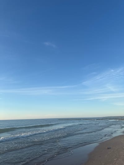 The image shows a view of the Baltic Sea coast in Ustka, Poland. The sky is a clear blue with some wispy white clouds. The sea is calm with gentle waves breaking on the sandy shore. In the distance, there are wooden groynes extending into the water. The beach is made of light brown sand. There are no people or animals visible in the image. The overall scene is serene and peaceful.
