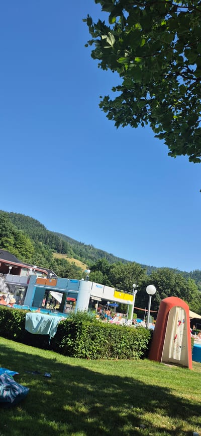 The image depicts an outdoor swimming pool complex on a sunny day, nestled against a backdrop of lush green hills. In the foreground, a well-maintained green lawn is visible, with some shadows cast from trees. To the right, a red, curved structure with a white shower curtain featuring a graphic design stands prominently. A thick, green hedge runs across the middle ground, separating the lawn from the pool area.
Behind the hedge, the swimming pool itself is visible, with several people swimming or relaxing in and around it. There are lounge chairs and umbrellas set up for sunbathing. Adjacent to the pool is a modern-looking building with a light-colored facade, featuring open sections and colorful accents of orange and yellow. This building appears to be part of the pool's facilities, possibly housing changing rooms or concessions.
Further in the background, more trees and vegetation cover the slopes of the hills, indicating a natural, somewhat secluded setting. The sky is a clear, vibrant blue. The architecture of the pool building and its amenities suggests a public leisure facility. Given the location described in the prompt, this scene is likely at the **"Freibad Forbach" (Forbach Outdoor Pool)**, a public swimming pool in Forbach, Germany, known for its scenic location in the Black Forest.