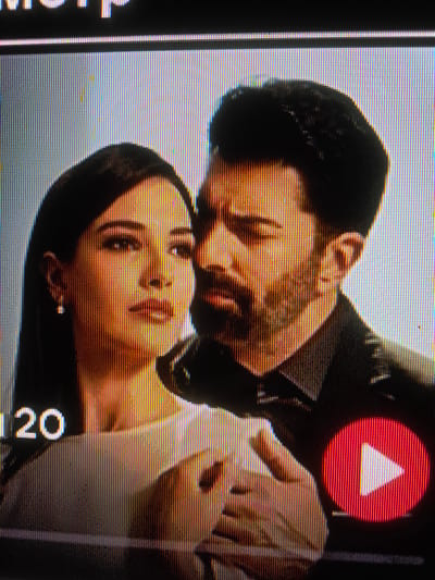 The image shows a close-up of two individuals, a woman and a man, in what appears to be a screenshot from a video. The woman is on the left, her face partially visible and turned slightly to the right, with dark hair framing her face. She is wearing a light-colored top and a small earring. The man is on the right, his face also partially visible, turned towards the woman. He has dark hair and a beard, and is wearing a dark shirt. In the lower left corner of the image, the number "120" is visible in white text against a dark background. In the lower right corner, there is a red circular play button with a white triangle inside, indicating a video playback interface. The background is a gradient of light and dark, with the overall impression being of a scene from a dramatic or romantic context. The resolution of the image is low, suggesting it might be a capture from a screen or a low-quality digital photo. There is no discernible information within the image to indicate the city of Shelkovskaya, Russia.