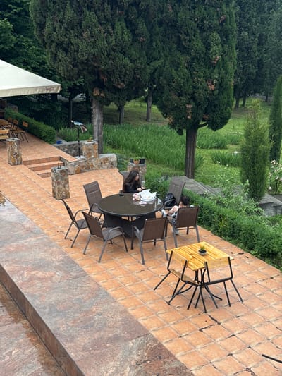 The image shows an outdoor seating area in Akhtala, Armenia, likely a restaurant or cafe, with a focus on a patio paved with terracotta tiles. In the mid-ground, a round, dark-colored wicker table is set with chairs, and two individuals are seated at it. One person, a woman with long dark hair, is leaning forward on the table, appearing to be eating or interacting with items on the table. The other individual, also a woman with dark hair, is seated with her legs crossed, looking away from the table. A black backpack is visible on the ground near her. To the right, a smaller, light-colored wooden table with two chairs is set, with a single small plant in a pot on its surface. In the background, lush green grass and mature cypress trees create a natural setting. To the left, a white awning with the Coca-Cola logo is visible, attached to a building structure. A stone staircase leads up to a slightly elevated area with more stone pillars and bushes. The ground near the foreground appears wet, suggesting recent rain.