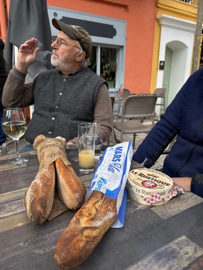 The image depicts an outdoor cafe scene in Cucuron, France. A middle-aged man with a white beard and round spectacles, wearing a brown cap, a brown long-sleeved shirt, and a dark grey button-up vest, is seated at a dark wood-grain patterned table. He is actively smoking a cigarette, with smoke visible exhaling from his mouth, and his right hand is raised, holding the cigarette.
On the table in the foreground, various items are arranged:
* A stemmed glass containing white wine.
* A tall clear glass holding a small amount of yellow liquid (juice).
* Another empty clear glass/pitcher.
* Two baguettes wrapped together in brown paper, labeled "Boulangerie Patisserie Gélis."
* A single baguette partially unwrapped from a blue and white paper bag, marked "KARS Cola" and "Vädops."
* A round wooden box of "LE RUSTIQUE Camembert" cheese, with a red and white checkered cloth visible beneath it.
To the right, a person's left arm, clad in a dark blue fleece-like jacket, rests on the table. In the background, an orange-red building facade with a grey-framed window is visible directly behind the smoking man, suggesting a cafe establishment. Further to the right in the background, a yellow building with a white arched doorway can be seen, typical of European town architecture. Several empty outdoor cafe tables and chairs are visible in the mid-ground, indicating a public dining area or square.