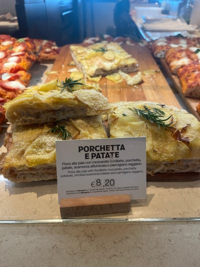 This image captures a close-up of a food display counter, likely in a bakery, pizzeria, or deli in Dorno, Italy. The central focus is a rectangular pizza, cut into several slices, prominently featured on a wooden cutting board. This pizza, labeled "PORCHETTA E PATATE," is topped with thin, lightly browned potato slices, visible chunks of porchetta, fresh sprigs of rosemary, and a sprinkling of grated cheese.
To the bottom left and in the background, other rectangular pizza slices are partially visible, one featuring a red sauce base with melted white cheese, suggesting a classic margherita or similar variety. The food is displayed under bright, likely artificial, lighting.
A white informational card stands next to the potato and porchetta pizza. The text on the card describes the dish in both Italian and English: "PORCHETTA E PATATE," "Pizza alla pala con mozzarella fiordilatte, porchetta, patate, scamorza affumicata e parmigiano reggiano" and "Pizza alla pala with fiordilatte mozzarella, porchetta, potatoes, smoked scamorza cheese and parmigiano reggiano." The price displayed is "€ 8,20." Additional text indicates that ingredients and allergens are listed on ingredient signs, and that the product's base was frozen at source.
No people are visible in the frame. The setting appears clean and organized, with the pizzas presented appealingly for sale. The time of day is not explicitly indicated but the bright lighting suggests daytime business hours.