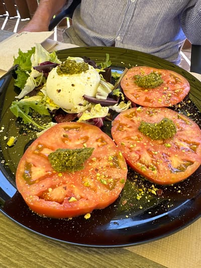 A close-up, overhead shot captures a dish of burrata cheese and sliced tomatoes with pesto, served on a black plate. The burrata, a ball of fresh mozzarella with a creamy center, is placed on a bed of mixed greens, including red leaf lettuce and arugula, and is topped with a dollop of vibrant green pesto. Three thick slices of ripe, red tomato are arranged on the plate, each adorned with a spoonful of pesto and sprinkled with chopped pistachios. The plate itself is speckled with tiny white dots, and some chopped pistachios are scattered across the black surface. The dish is presented on a textured, wooden table, with a person's arm and shoulder, clad in a blue and white striped shirt, visible in the upper background, suggesting a dining setting. The lighting suggests an outdoor or well-lit indoor environment. This dish is likely from a restaurant in Santa Cristina d'Aro, Spain, known for its fresh Mediterranean cuisine.