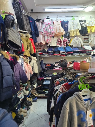 The image displays the interior of a retail clothing store located in Taza, Morocco. The space is densely merchandised with garments and footwear, primarily aimed at children and young adults. The left side of the frame features multiple rows of hanging jackets and vests in various colors (e.g., navy, gray, olive, red, purple). Beneath these, several pairs of shoes, including dress shoes and boots, are arranged on lower shelves. The back wall showcases a selection of children's clothing, including hanging dresses (one white with pink floral patterns, one peach) and various tops (e.g., a black skeleton print shirt, a white striped cardigan). Folded stacks of clothing items are neatly organized on shelves across the back. In the right foreground, a metal rack holds additional garments, including patterned sweaters and a gray hooded sweatshirt with a blue "23" graphic, bearing a "Seramis" brand tag. The store has white walls, a white tiled floor with a diamond pattern, and is illuminated by overhead fluorescent strip lights and a circular ceiling light. A black dome-shaped security camera is mounted on the ceiling in the upper left. No individuals are present in the scene.