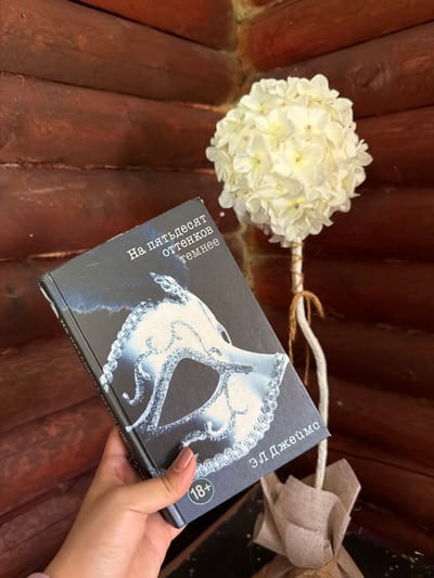 A hand with a light pink nail polish holds a book titled "Fifty Shades Darker" in Russian, with a masquerade mask as the cover image. The book is placed against a dark wooden log wall. To the right of the book, a white hydrangea flower arrangement is displayed on a stand, tied with twine and burlap fabric. An "18+" symbol is visible on the bottom right corner of the book cover. The lighting suggests an indoor environment with warm tones. The scene does not contain elements that can identify a specific location in Nagornyi, Russia.