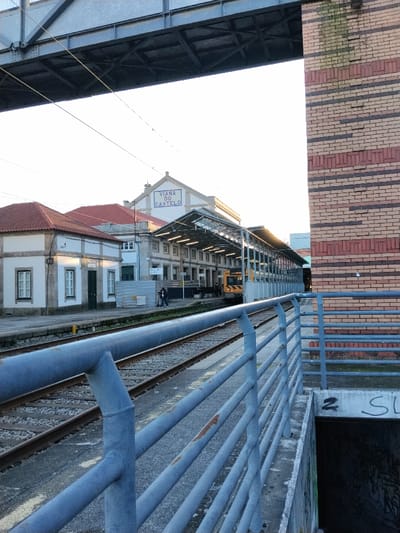 A train station platform is viewed from behind a blue metal railing. Railway tracks run parallel to the platform. Buildings, including the "VIANA DO CASTELO" station, line the opposite side of the tracks. A yellow train sits at the platform. Part of a bridge structure is visible overhead. To the right, a brick building with horizontal red, brown, and black stripes abuts the platform. Graffiti is visible near an underground entrance.