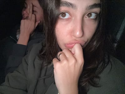 A selfie of a person with dark hair and brown eyes is oriented upside down, with a hand on the upper left side of the image pulling on the person's lip. A gold ring is visible on the hand. Another person’s face is partially visible on the lower right, with a hand near their face. The background is dark and indistinct. There is no indication of the city of Puerto Marqués, Mexico, in this image.