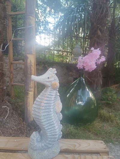 This is a medium shot, eye-level, outdoor photograph taken in Serravalle, Switzerland. The foreground features a light grey, concrete statue of a seahorse, positioned on a light-colored wooden pallet. The seahorse statue has textured detailing and a seashell accent on its side. To the left of the seahorse is a tall, bamboo pole structure with horizontal wooden rungs, resembling a ladder. Behind the seahorse and to the right, a large, dark green glass bottle is visible, containing a bouquet of small, pink flowers. In the background, there is dense foliage, including palm fronds, and a blurred, decorative metal fence. The lighting suggests it is daytime, with some lens flare from the sun. The environment appears to be a garden or an outdoor decorative space.