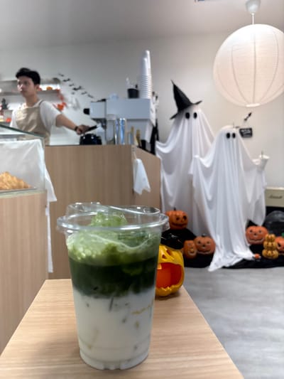 An interior view of a cafe in Tambon Hua Toei, Thailand, features Halloween decorations. In the immediate foreground, a clear plastic cup containing a layered iced beverage rests on a light wood-grain counter. The drink exhibits a white opaque layer at the bottom, a dark green layer above it, and a lighter green frothy layer with several green ice cubes on top. To the right of the cup, a small orange pumpkin-shaped lantern with a black conical hat is visible.

In the midground, a male individual with dark hair, wearing a white t-shirt and a light beige apron, stands behind a light wood-paneled counter, operating an espresso machine. White disposable cups are stacked on the counter to his right. Black bat cutouts adhere to the white wall behind him, and a display case with baked goods, possibly croissants, is partially visible to his left.

Further to the right, two ghost figures, fashioned from white sheets with black oval eyes, are positioned. The taller ghost wears a black witch's hat. They are surrounded by a collection of orange pumpkin-shaped decorations of various sizes, some with carved faces, placed on a dark surface. A large white spherical paper lantern hangs from the ceiling above this display. The cafe walls are white, and the floor appears to be a light gray concrete.