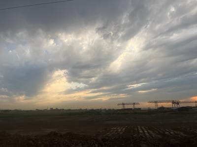 The image depicts an industrial landscape under a dramatic, cloudy sky. The foreground is dominated by dark, rough terrain, possibly construction or industrial waste, with visible tire tracks in the mud. In the mid-ground, a vast, flat expanse of land stretches towards the horizon, dotted with various industrial structures. Most prominent are several large, yellow gantry cranes, silhouetted against the sky, suggesting ongoing construction or manufacturing operations. Scattered among the cranes are smaller buildings and structures, hinting at a developed industrial zone. The background features a hazy horizon with more distant structures and possibly mountains or hills obscured by atmospheric conditions.
The sky is filled with heavy, dark clouds, but patches of bright, pale yellow light break through, particularly on the left and right sides of the image, indicating either sunrise or sunset. This contrast between the dark, imposing clouds and the illuminated sky creates a sense of drama and atmosphere. The overall mood is somewhat somber and industrial, yet the light breaking through suggests a glimmer of hope or the transition of time. The location is Pervomayskiye Prudy, Kazakhstan. There is no visible text in the image. The time of day is likely early morning or late evening due to the lighting conditions. The weather appears overcast with potential for rain.