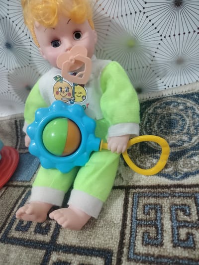 A doll with blond hair, blue eyes, and a pacifier is sitting on a patterned rug. The doll is wearing a light green outfit with a white collar and cuffs. It is holding a blue rattle with a green and orange ball inside. The background is a white surface with a geometric pattern. The location is the city of Cholpon, Kyrgyzstan.