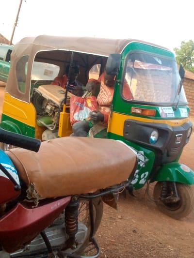 A three-wheeled motorized vehicle, painted green and yellow, has two occupants in the back. A generator sits next to them. The passenger in front holds a bag labeled "Veep." The vehicle has "RE" on its front panel and a design featuring white numbers on its side. In the foreground, a motorcycle with a brown seat is visible. The surrounding environment includes a red dirt surface and blurred buildings in the background.