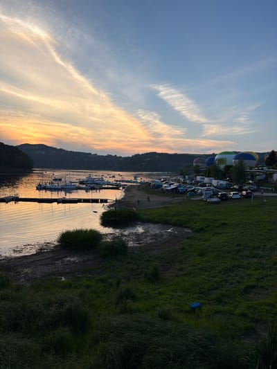 This is a vertical, medium-shot, eye-level, full-color photograph taken at sunset in Gródek nad Dunajcem, Poland, at Lake Rożnowskie. The sky is a gradient of light blue and orange, with wispy clouds. The lake is calm, reflecting the colors of the sky. Several sailboats are docked along the shore, and a few people are visible walking on the beach. In the background, there are hills covered with trees. Further inland, there is a grassy area with several parked cars and camper vans, and a few large, colorful hot air balloons are inflated and tethered. The overall scene depicts a peaceful evening by the lake, with recreational activities and a hot air balloon festival.
