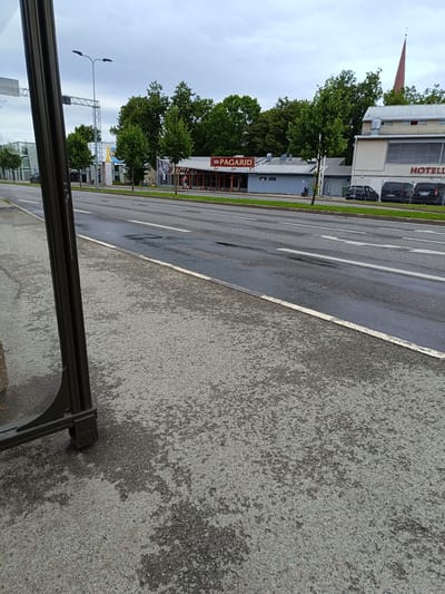 This is an eye-level, medium shot of a street scene in Jõhvi, Estonia. The foreground is dominated by a dark gray, rough-textured concrete sidewalk and a dark gray metal pole, part of what appears to be a bus stop shelter. The sidewalk has patches of darker wetness, suggesting recent rain. Beyond the sidewalk is a wet asphalt road with white lane markings. In the middle ground, on the opposite side of the street, are several buildings and trees. A building with a red sign reading "PAGARID" (Estonian for "bakers") is visible, along with a larger building to its right with a sign that reads "HOTELL" (Estonian for "hotel") and a tall, slender, reddish-brown spire of a church or steeple is visible behind the hotel. Several vehicles, including black vans and cars, are parked in front of the hotel. The background consists of a dense line of green trees and an overcast, cloudy sky. The overall atmosphere is somewhat somber due to the cloudy weather and wet conditions. There are no prominent individuals or specific actions captured in this frame.