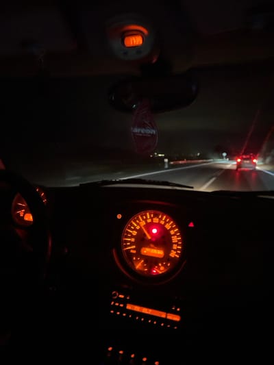 The image captures the interior of a vehicle at night, likely in Kujava, Montenegro, with a focus on the dashboard and part of the windshield view. The dominant feature is the illuminated dashboard, particularly the large central speedometer, glowing in orange and red. The speedometer indicates speeds up to 240, presumably km/h, and shows a digital odometer reading of "999222.8". A red warning light is illuminated on the speedometer face. To the right of the speedometer, part of the center console with various illuminated orange controls and buttons is visible.
On the left side of the frame, near what appears to be the ignition or a start button, a small digital display shows "1733", next to which a "SET" button is visible. A red air freshener, branded "areon", hangs within the cabin. Through the dark windshield, the faint red taillights of another vehicle are visible in the distance, along with some indistinct lights of a road or environment, confirming the nighttime setting. The vehicle interior is largely dark except for the dashboard lights, which provide the primary illumination for the scene. The overall impression is that of a car either driving or idling on a road at night.