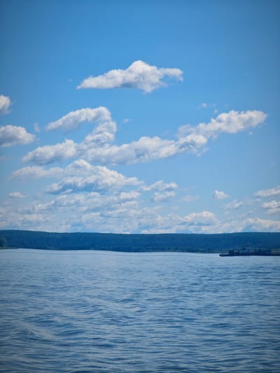 The image depicts a wide, calm body of water, likely a river, under a bright blue sky dotted with white, fluffy clouds. A dark green, tree-covered shoreline stretches across the horizon. In the distance on the right side of the frame, a small tugboat is visible moving across the water. The scene is tranquil and suggests a clear, pleasant day. The location is identified as Krutoi Log, Russia.