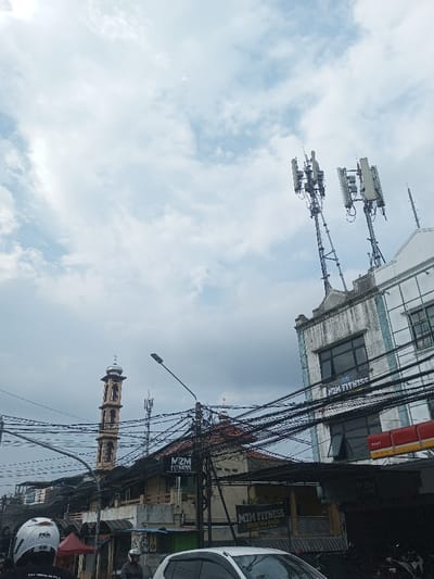The image shows an urban scene with a tall minaret and cell towers. Overlapping power lines obscure the view of the building's exterior and the street below. The sky is cloudy, suggesting overcast weather. A white car and people on motorcycles are visible in the foreground, indicating street-level activity. The buildings exhibit a mix of old and modern architecture. "M2M FITNESS" is visible on some buildings.