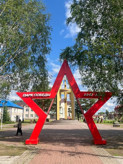This is a vertical, eye-level, outdoor photograph taken in Mezhdurechenskiy, Russia. The image features a large, bright red, abstract sculpture with angular geometric shapes, set against a backdrop of trees with green foliage and a bright blue sky with scattered white clouds. The sculpture is in the foreground, dominating the center of the frame. It appears to be made of metal and has two distinct red angular elements that meet at a central point, forming an arrowhead-like shape.
Visible text on the sculpture reads "1941-1945" on one of its angled surfaces. On another surface, oriented vertically, the Cyrillic text reads "ПАРК ПОБЕДЫ" which translates to "Victory Park".
In the background, beyond the sculpture, there is a paved walkway or plaza made of red bricks. Further back, there are buildings with light-colored facades and pitched roofs, suggesting a residential or public area. The presence of trees and the blue sky with clouds indicate it is daytime, likely pleasant weather.
A small figure of a person can be seen in the lower right portion of the image, walking on the paved area. The overall impression is one of a public space, possibly a memorial or entrance to a park, marked by the prominent red sculpture commemorating a historical period. The bright colors and clear sky suggest a cheerful or celebratory atmosphere, despite the memorial inscription.