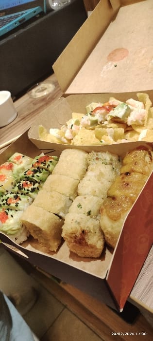 The scene depicts an open brown cardboard takeout box containing multiple food items, positioned on a light-colored wooden surface. In the upper compartment, tortilla chips are topped with white sauce, diced green components (likely avocado or cucumber), and pink shrimp pieces. The lower compartment contains four distinct rows of sushi-style rolls: a left-most row with green-wrapped rolls topped with black sesame seeds and red roe; two central rows of light-colored, possibly tempura-fried or breaded rolls, one with visible green specks; and a right-most row of rolls featuring a glossy, brownish-orange topping. No individuals are visible. In the background, a dark electronic device, likely a laptop, with a partial light blue keyboard is visible, alongside a white cup. The environment is an indoor setting, likely a home or restaurant, in Tumbaco, Ecuador. A timestamp of "24/02/2026 17:38" is present in the lower right corner. No actions or interactions are currently occurring within the frame.