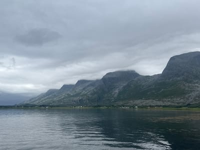 This media file depicts a serene, cloudy landscape scene in Sandnessjøen, Norway. The foreground is dominated by a wide expanse of calm water, reflecting the overcast sky and the mountainous terrain beyond. Gentle ripples disturb the surface of the water, suggesting a light breeze.
In the middle ground, a low-lying shoreline is visible, dotted with a scattering of small houses and buildings, indicating a coastal village or settlement. Beyond the settlement, steep, rugged mountains rise dramatically, their peaks partially obscured by thick, grey clouds. The mountains have a textured appearance, suggesting rocky slopes with sparse vegetation. Some of the higher elevations are shrouded in mist, contributing to the moody atmosphere.
The sky is a uniform, muted grey, indicative of overcast weather, possibly with rain or fog in the distance, particularly on the far left where the sky appears darker. The overall lighting is diffused, creating a soft, subdued ambiance. There is no visible text within the image. The scene conveys a sense of peaceful solitude and the raw beauty of a northern coastal environment under a cloudy sky. The time of day is not explicitly discernible but appears to be daytime, given the level of ambient light.
