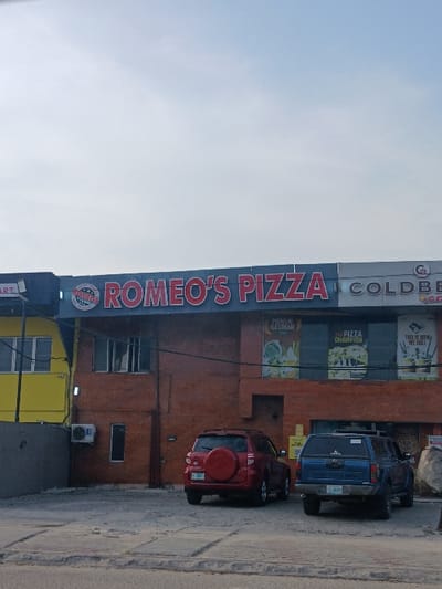 This image displays the exterior of a commercial building in Ojodu, Nigeria, under an overcast sky. The building features a large sign reading "ROMEO'S PIZZA" in red capital letters on a dark background, with a circular logo to its left. To the right of the pizza sign, a partially visible sign reads "COLDBE_ _ _" with a 'G' logo. The ground floor of the building is constructed with reddish-brown brickwork and displays several large promotional posters, including one for "PREMIUM ICE CREAM" and another stating "BE A PIZZA CHAMPION." An external air conditioning unit is mounted on the wall to the left on the ground floor. A bright yellow building is partially visible to the left, adjacent to the main structure. Two vehicles are parked on the paved forecourt directly in front of the building: a red SUV, likely a Toyota RAV4, with a red spare tire cover mounted on its rear door, and a blue pickup truck equipped with a bed cap and a roof rack, displaying a Lagos state license plate (TFK-053XA). No individuals are visible in the scene, and no actions or interactions are depicted.