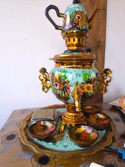 A brightly colored, ornate samovar is the primary subject of this image. It is made of polished brass and decorated with vibrant floral patterns in blues, reds, yellows, and greens. The samovar features an upward-curving handle and a spigot. It sits on a decorative wooden tray that is also embellished with floral motifs, this time in silver and black. Several small, lidded bowls, similarly decorated with red berries and gold leaves, are arranged on the tray. The background consists of a white wall and a wooden surface, suggesting an indoor setting. The lighting appears to be artificial and even. No people are visible in the image. The overall impression is one of traditional craftsmanship and decorative artistry. The location context is Reisbach, Germany. There is some blurry text visible at the top of the image, but it is not legible.