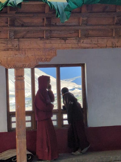 In Yulang, India, two individuals dressed in traditional maroon robes are observed standing near a large window, gazing out at a mountainous landscape. The person on the left, with a hood pulled up, appears to be holding their hand to their face, possibly in thought or conversation. The individual on the right leans against the window frame, looking out. The window is divided into smaller panes and offers a view of a valley with buildings and a river. The interior features a wooden beam overhead and a dark red baseboard along the wall.