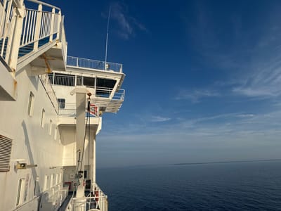An eye-level, wide shot captures the side of a white ferry boat sailing on a calm, blue sea under a bright, clear sky. The ferry's white structure dominates the left side of the frame, featuring multiple levels, white railings, windows, and exterior staircases. A prominent mast extends upwards from the top deck. The sea stretches across the majority of the frame to the right, meeting a hazy horizon. A faint, distant landmass is visible on the horizon. The sky is a deep blue, with scattered wispy white clouds. No people are visible in the image. The scene is a daytime, outdoor shot, likely taken from another vessel or the deck of the ferry itself. The location is not specified as being within a city in Sweden.