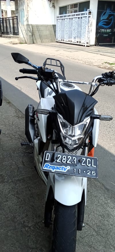 A white Honda motorcycle with black accents is parked on a paved street in Ciapus, Indonesia. The motorcycle is facing the viewer and is the primary subject. It features a black front fender, a black windscreen, and a black headlight assembly with clear lenses. The handlebars are silver with black grips and mirrors. A black metal rack is mounted behind the handlebars. The motorcycle has a license plate that reads "D 2823 ZCL" and "Rzqactv 11-26". The motorcycle is partially obscured by a gray concrete curb and sidewalk. In the background, there is a building with a white facade and a large, decorative white gate. A blue logo with a fox silhouette is visible on the side of the building. The scene appears to be taken during daylight.