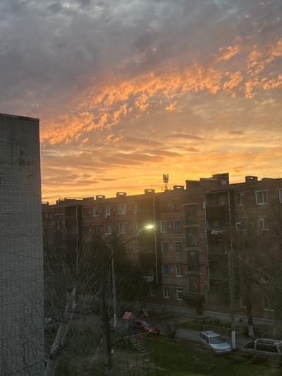 The image depicts an urban residential area in Dmitrievskaya, Russia, at either sunrise or sunset. The sky displays a dramatic gradient, transitioning from grey-blue in the upper left to vibrant orange and yellow hues across the mid-section, illuminating scattered clouds. A partial view of a tall, grey brick building occupies the left foreground. In the midground, a multi-story, reddish-brown brick apartment building extends horizontally, featuring numerous windows, multiple balconies (some enclosed), and visible roof structures including a cellular telecommunications antenna array. A bright streetlamp, emitting a yellowish-green light, is visible in the lower left-center, illuminating parts of the building facade and the street below. At ground level, several bare deciduous trees with visible branches are present. A paved street or parking area contains three parked vehicles: a silver station wagon, a darker station wagon, and a partially visible red car. A red-framed playground structure is visible in the lower left corner. No individuals or discernible activity are present within the scene.