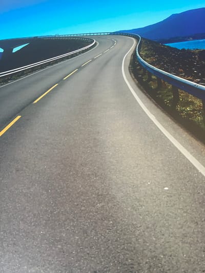 This is a photograph of a winding road with guardrails, bordered by trees and a body of water. The road has white and yellow lane markings. The sky is a clear, bright blue. The overall scene appears to be a scenic mountain or coastal road. The image is taken from a high angle, looking down at the road as it curves away. There are no people visible in the image. The lighting suggests it is daytime, likely with fair weather.