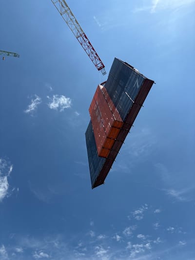 In Cheongju-si, South Korea, a construction site is active under a clear blue sky with scattered white clouds. A large red and white crane arm extends from the upper left, supporting a suspended construction module. This module, appearing to be part of a building's facade, is rectangular and multifaceted, with sections of dark mesh and orange material. The orange sections have white text, partially visible, including the number "1" and what appears to be the letters "ONM". The module is suspended at an angle, suggesting it is in the process of being lifted or positioned. In the far upper left, another, smaller green crane is visible. The perspective is from below, looking directly up at the sky and the construction elements.