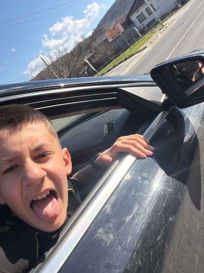 A young person is sticking their tongue out of a car window. Their hand is visible, gripping the car door frame. The car appears to be in motion on a road near the city of Zlatna Panega, Bulgaria, as suggested by the signage visible in the background. The sky is bright blue with sparse clouds.