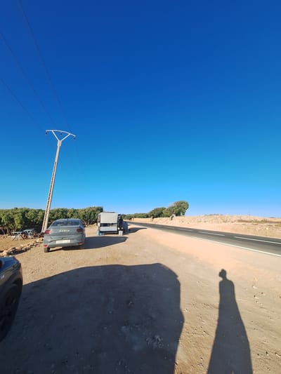 This is a landscape photograph taken outdoors on a bright, sunny day, likely in the afternoon given the long shadows cast. The scene depicts a rural roadside in Ahrara, Morocco. A paved road with white lane markings runs diagonally through the frame, bordered by a dusty, arid ground with scattered rocks and vegetation.

The primary subjects in the image are vehicles and infrastructure. A dark grey Dacia car is parked on the side of the road, facing away from the viewer. Behind it, a trailer with a gray cover is attached to another vehicle, which is partially visible. In the distance, more vehicles and trailers are present. A prominent utility pole with power lines stretches across the sky, indicating the presence of electrical infrastructure in the area. The background is a clear, vibrant blue sky, suggesting fair weather with no clouds.

A notable detail is the shadow of a person, likely the photographer, cast on the ground, stretching from the right side of the image. This provides a sense of scale and confirms the presence of human activity. The overall atmosphere is one of a quiet, rural setting, possibly a stop along a journey.

Visible text on the Dacia car's license plate reads "50901 154". The word "DACIA" is also visible on the rear of the car. The dry, rocky terrain and sparse vegetation are consistent with the arid climate often found in Morocco.