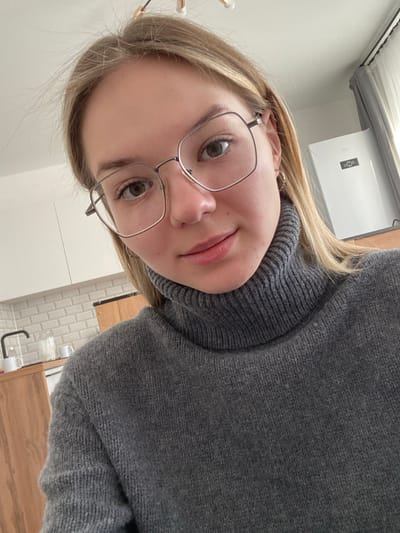 An image taken in a residential interior, specifically a kitchen area, in the city of Bittsa, Russia, depicts a single female subject in a close-up, likely a selfie. The subject, positioned centrally, has fair skin, brown eyes, and blonde hair, which is parted and extends over her shoulders. She wears large, silver-framed, hexagonal eyeglasses and a dark grey knit turtleneck sweater. A small silver hoop earring is visible in her right ear. The subject is looking directly towards the camera with a neutral to slight smiling expression.
The background shows a contemporary kitchen design. To the subject's left, white upper cabinets, a white subway tile backsplash, and light brown wooden lower cabinets or countertop are visible. A black gooseneck faucet, white mugs, and a glass container are present on the counter. To the subject's right, a white appliance, possibly a refrigerator or air conditioning unit, with a small digital display is seen. Grey curtains or drapes are partially visible next to this appliance. Overhead, a modern light fixture with multiple light sources on gold-colored arms is partially visible on a white ceiling.