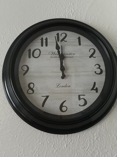 The image shows a close-up of a round, black-framed clock with a light wood-grain background. The clock face has large, dark numerals. The hour hand is pointing to the 12, and the minute hand is also pointing to the 12, indicating the time is exactly 12:00. The words "Westminster Clock Company" and "London" are visible on the clock face. The clock is mounted on a textured white wall. The location of the city is not determinable from the image.