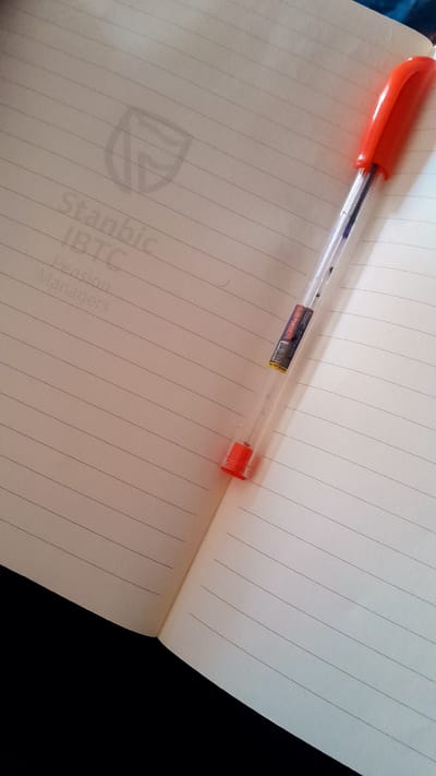 An orange and clear ballpoint pen rests diagonally across an open, ruled notebook, captured from a high-angle perspective. The pen's transparent barrel reveals a blue ink cartridge and a small white label with a barcode. Visible text on the pen's label includes "iTcraft" and "LEADER".
The notebook pages are lined with horizontal rules. The right page is largely blank, while the left page displays a faint, light grey watermark and text oriented vertically. The visible text on the notebook reads: "Stanbic", "IBTC", "Pension", and "Managers". The background around the notebook is dark, suggesting a shadowed surface. The scene is well-lit, likely indoors, with soft lighting. No active writing is occurring, but the arrangement suggests a workspace or a momentary pause from note-taking. The location context is Umuigbekwo, Nigeria.