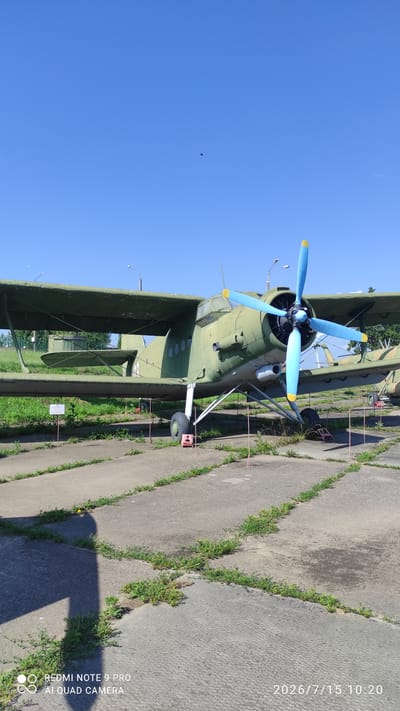 A green biplane with a blue propeller is parked on a concrete tarmac under a clear blue sky. The aircraft has the number "41" painted on its fuselage. The tarmac is cracked and overgrown with grass in places. Several orange markers are visible around the aircraft, and a white sign is posted to the left of the plane. In the distance, trees and a fence can be seen. The scene is illuminated by bright sunlight, casting a shadow of the aircraft onto the tarmac in the foreground. The date stamp "2026/7/15 10:20" and "REDMI NOTE 9 PRO AI QUAD CAMERA" are visible in the bottom right and left corners respectively.