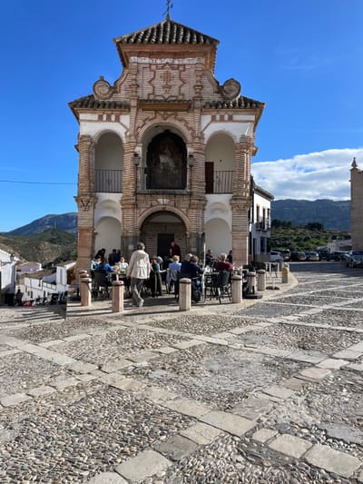 The image shows a view from the ground looking up at a stone building with several arches. There are people sitting at tables and chairs on the cobbled square in front of the building. The image is taken in the city of Antequera, Spain, located at 37.014 latitude and -4.56 longitude. The sun is shining in the sky and there are mountains in the background. The building is a historic structure and it is a popular place for tourists and locals alike. The square is a lively and busy area. The people are interacting with each other and enjoying the warm weather. The image has a sense of place and history, and it is a beautiful example of Spanish architecture.