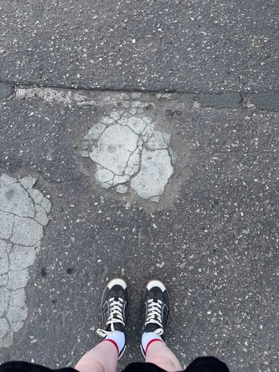 A high-angle shot depicts a section of cracked asphalt pavement. A repair patch, characterized by a lighter-colored and heavily fractured concrete-like material, is visible near the center. To the right of the frame, a pair of feet wearing black and white sneakers with white laces and red-striped white socks is present. The person wearing the shoes is also wearing a black garment. The location, Balezino, Russia, cannot be verified from this image.